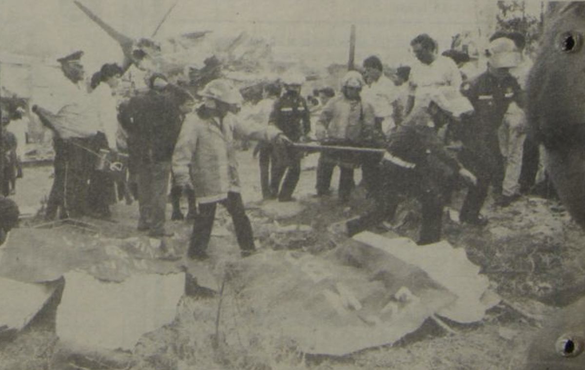 Los bomberos colocan varios cadáveres en un terreno baldío situado frente al lugar donde cayó el avión, el cual se observa al fondo, destruido. 5/5/1990. (Foto: Hemeroteca PL)