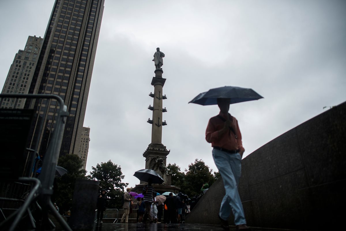 Varias personas camina cerca de la  la estatua de Cristóbal Colón en Nueva York mientras que un  grupo de personas pidela eliminación del monumento.