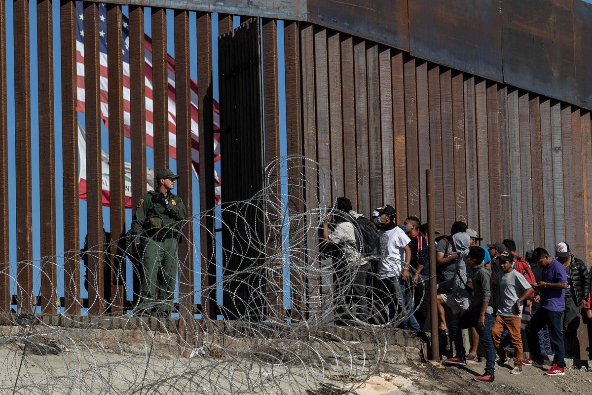 Migrantes centroamericanos miran a través de una valla fronteriza mientras un agente fronterizo de los EE. UU. hace guardia cerca del cruce fronterizo de El Chaparral en Tijuana. (Foto Prensa Libre: AFP)