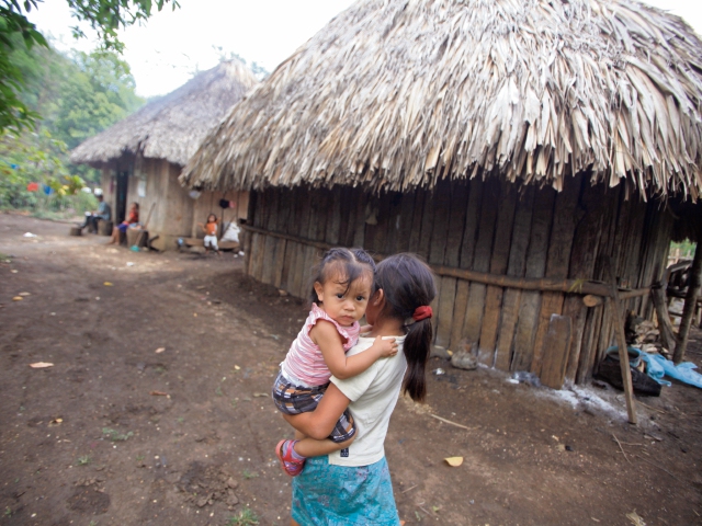 Una niña carga a su hermana menor mientras camina por la calle rural hacia su casa, en San Marcos, Poptún, Petén. Las viviendas son mínimas.