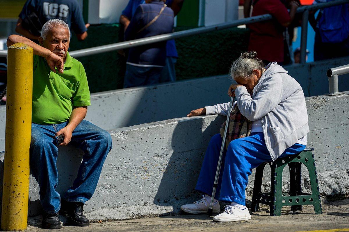 Dos personas de la tercera edad descansan afuera de un centro comercial mientras otras personas hacen fila para obtener alimentos en Caracas, Venezuela. (Foto Prensa Libre: AFP).