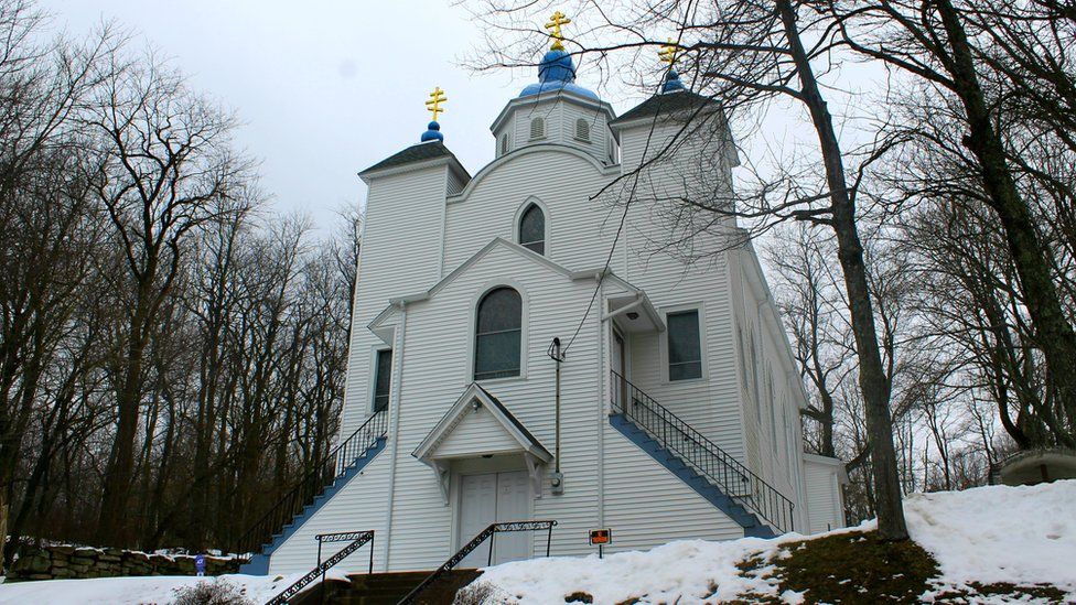 Una iglesia blanca se levanta entre árboles negros. Una cúpula azul brilla contra la nieve.