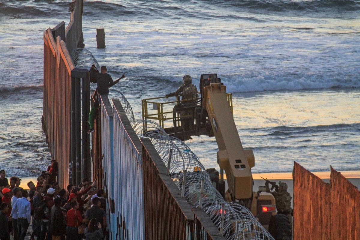 Unos migrantes trepan el muro fronterizo en Tijuana, México, mientras un soldado apunta su arma contra ellos del lado estadounidense. (Foto Prensa Libre: EFE)