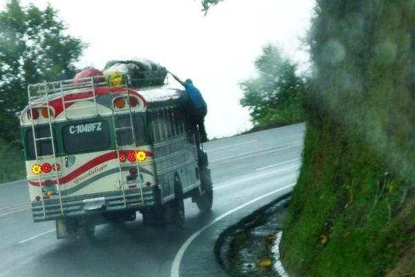 Esta foto fue tomada cerca de Sololá, carretera Interamericana. El ayudante del autobús extraurbano  amarra la carga y después baja a la puerta, mientras el vehículo viaja a una velocidad aproximada de 80 kilómetros por hora.