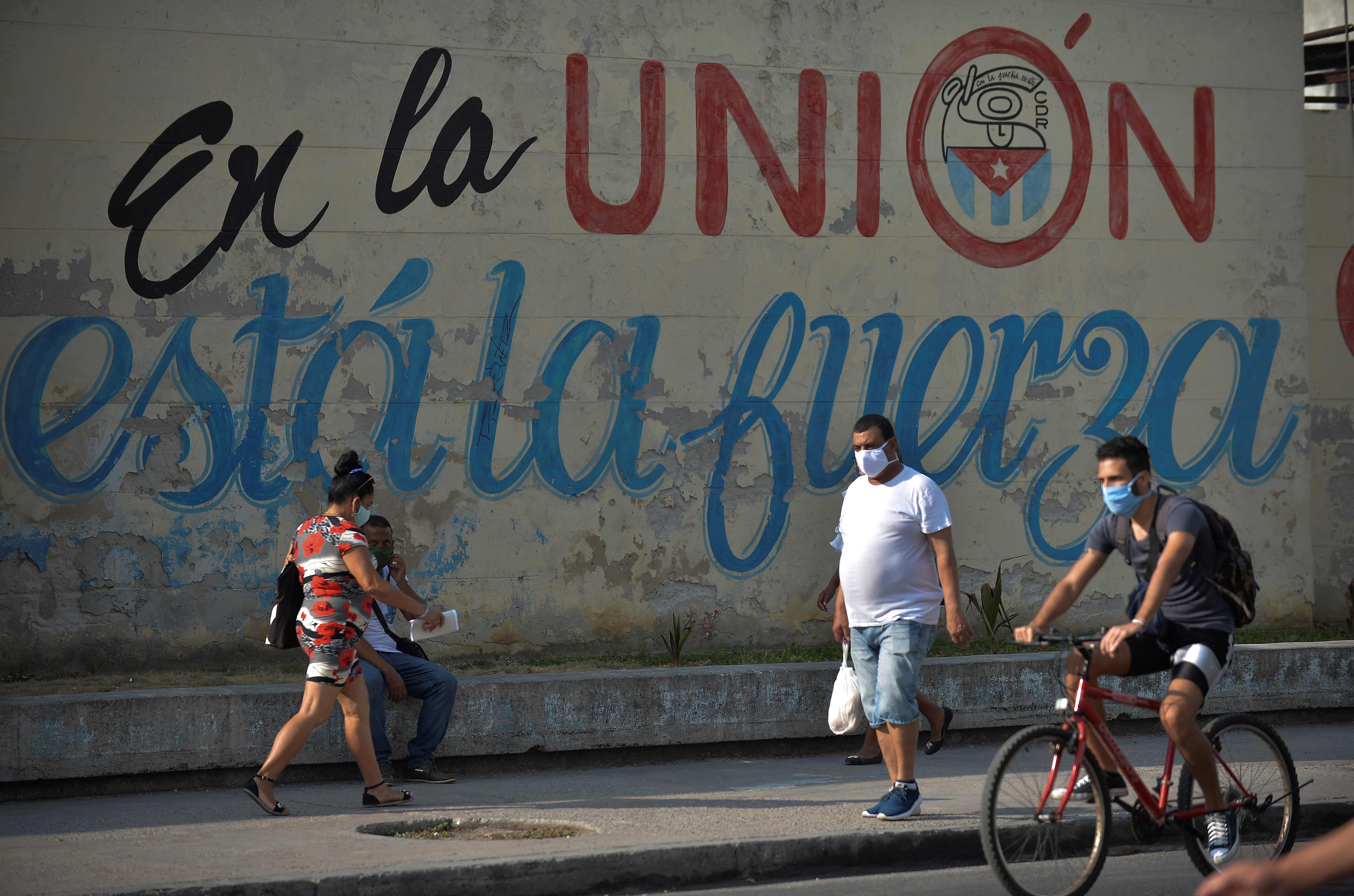 People wear face masks near a wall reading Union makes force in a street of Havana, on April 13, 2020. - While Cuba tries to stop the spread of the coronavirus on the island, with 726 cases, voices can be heard to demand the lifting of the US embargo to facilitate the arrival of medical products. (Photo by YAMIL LAGE / AFP)