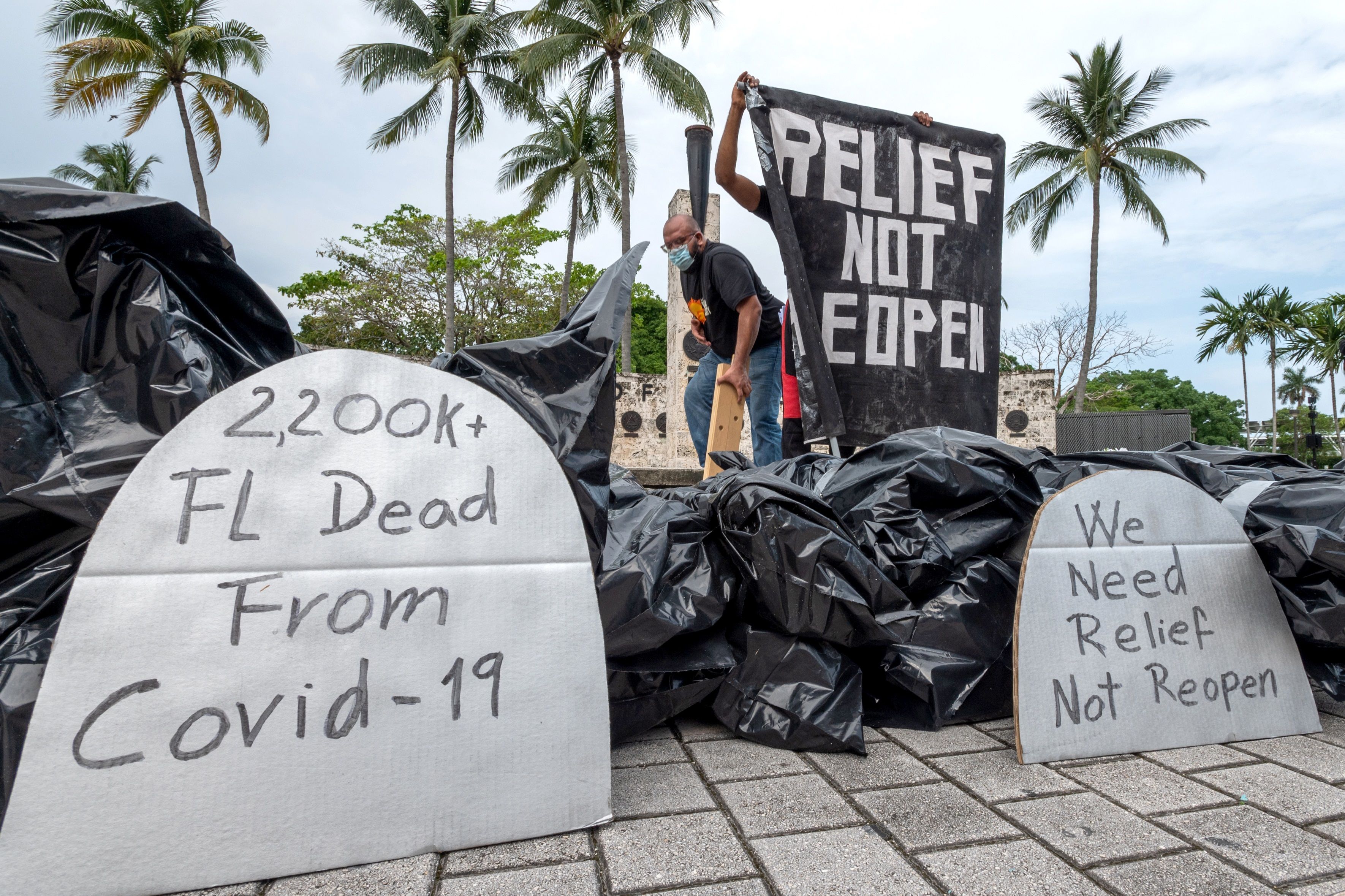 Una persona despliega una pancartas frente a unas bolsas con supuestos "cadáveres" que representan a los muertos por coronavirus este 27 de mayo durante la protesta contra la reapertura de la economía en el parque de la Antorcha de la Amistad en el centro de Miami, Florida. (Foto Prensa Libre: EFE).