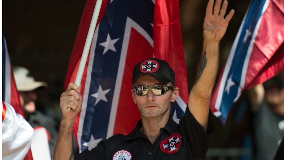 Un supremacista hace el saludo nazi con una bandera de la confederación y las cruces símbolo del Ku Klux Klan.
