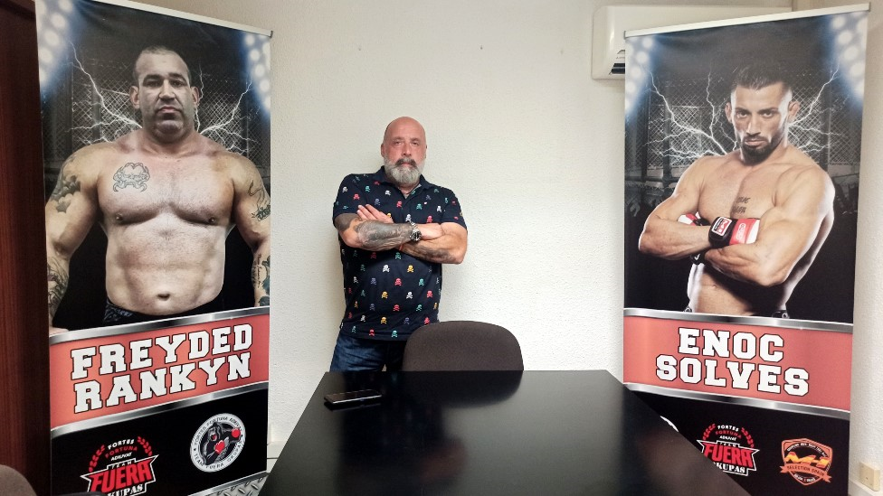 Jorge Fe posa entre la foto de dos de sus empleados que trabajan amedrantando okupas para recuperar propiedades. BBC