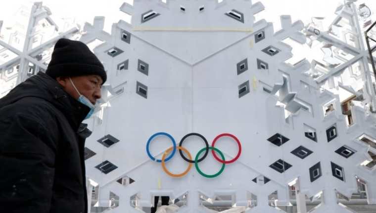 Un hombre con mascarilla y abrigo camina frente a un edificio con el logo olímpico.