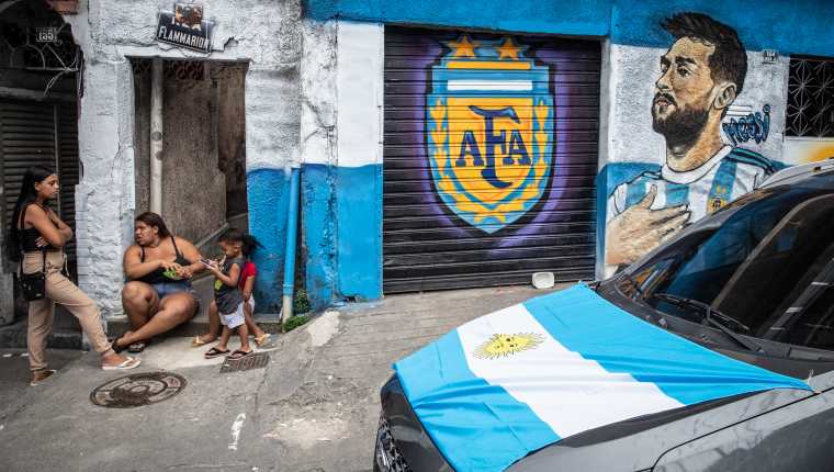 Vecinos conversan frente a sus casas pintadas con el símbolo de la Asociación del Fútbol Argentino (AFA), la imagen del jugador Lionel Messi y un auto con la bandera argentina. (Foto Prensa Libre: EFE)