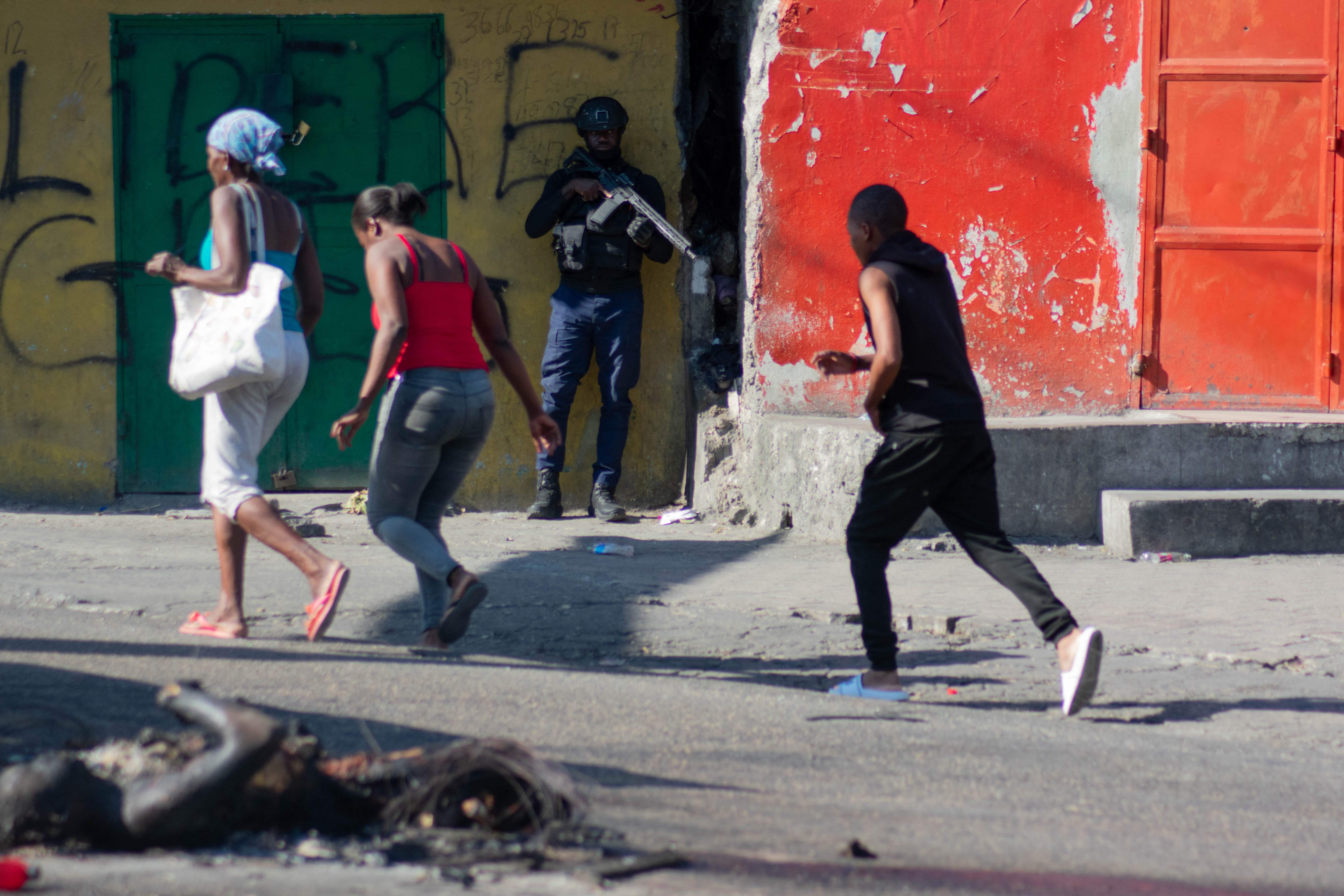 EDITORS NOTE: Graphic content / People walk past burned bodies and an armed police officer monitoring a street after gang violence in the neighborhood on the evening of March 21, 2024, in Port-au-Prince, Haiti, March 22, 2024. More than 33,000 people fled Port-au-Prince this month as the Haitian capital was overrun by well-armed gangs triggering political chaos in the impoverished Caribbean nation, the United Nations has said. (Photo by Clarens SIFFROY / AFP)