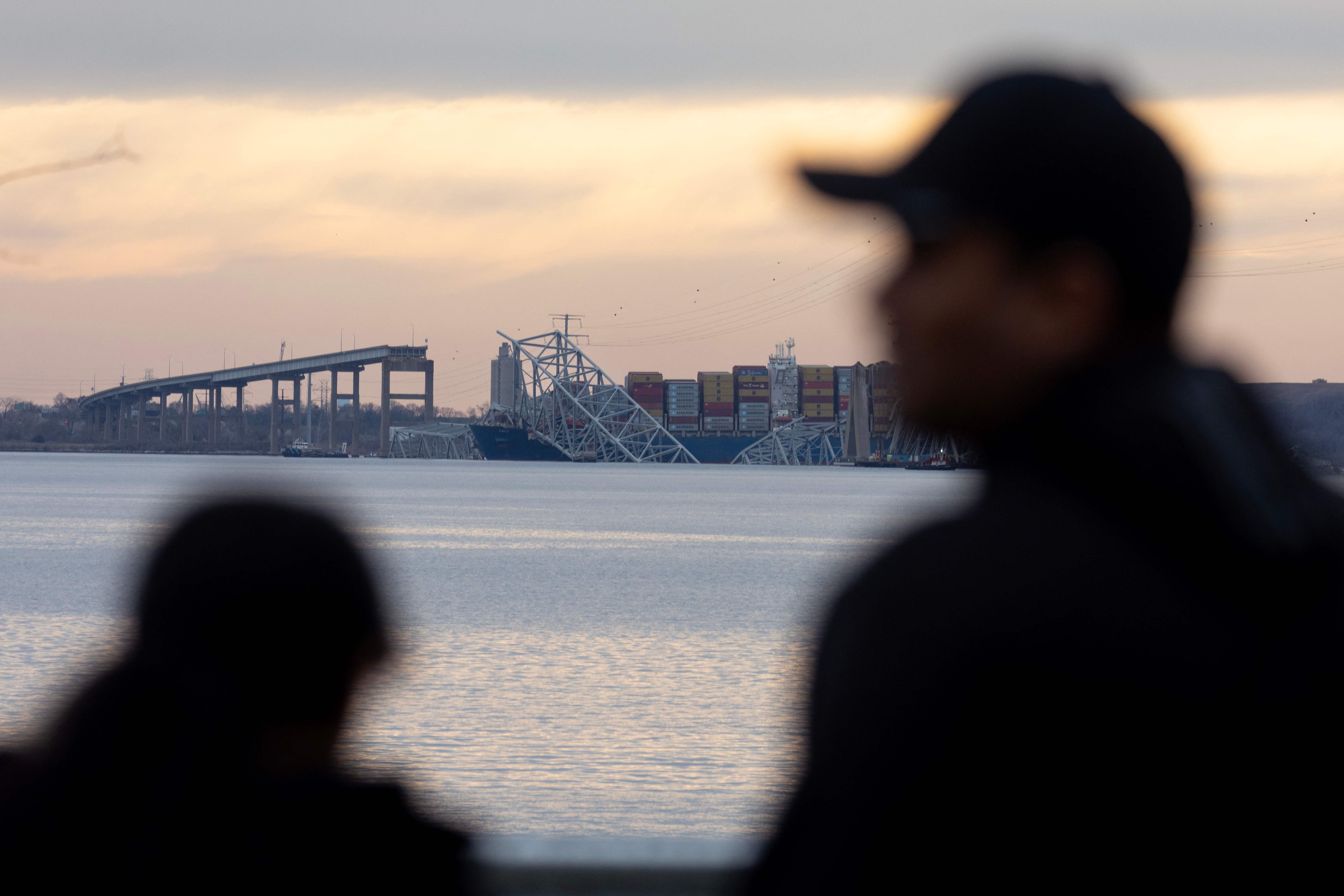 BALTIMORE, MARYLAND - MARCH 28: People watch as the sun descends beyond the collapsed Francis Scott Key Bridge on March 28, 2024 in Baltimore, Maryland. The bridge, used by roughly 30,000 vehicles each day, fell into the Patapsco River after being struck by the Dali, a cargo ship leaving the Port of Baltimore at around 1:30 a.m. Tuesday morning. The bodies of two men who were on the bridge at the time of the accident have been recovered from the water, with four others still missing and presumed dead and two others rescued and treated for injuries shortly after the accident. The Port of Baltimore, one of the largest and busiest on the East Coast of the U.S., is temporarily closed.   Scott Olson/Getty Images/AFP (Photo by SCOTT OLSON / GETTY IMAGES NORTH AMERICA / Getty Images via AFP)