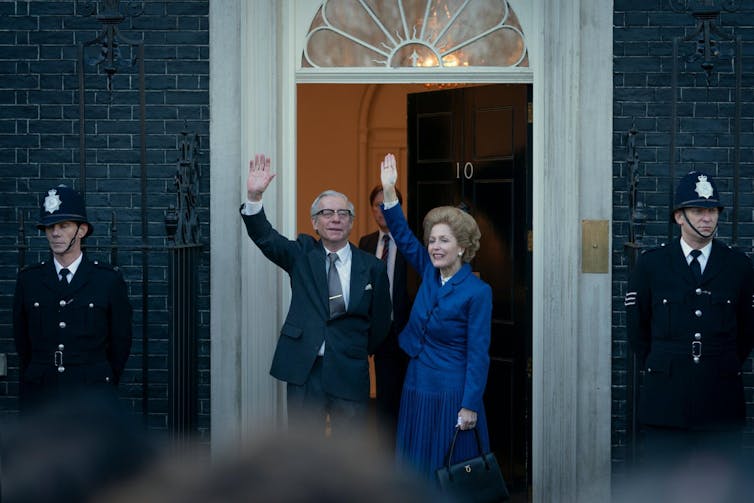 Una mujer con traje azul saluda a la puerta de una casa al lado de un hombre.