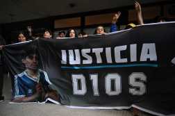 Fans gesture as they hold a flag with his image of late Diego Maradona in front of the San Isidro court ahead of the trial for the death of late Argentine football legend Diego Maradona in San Isidro, Buenos Aires province, Argentina on March 11, 2025. Four years after Diego Maradona's death, the trial of seven health professionals begins this Tuesday in Argentina to determine their responsibilities in the death of the soccer legend. (Photo by Luis ROBAYO / AFP)