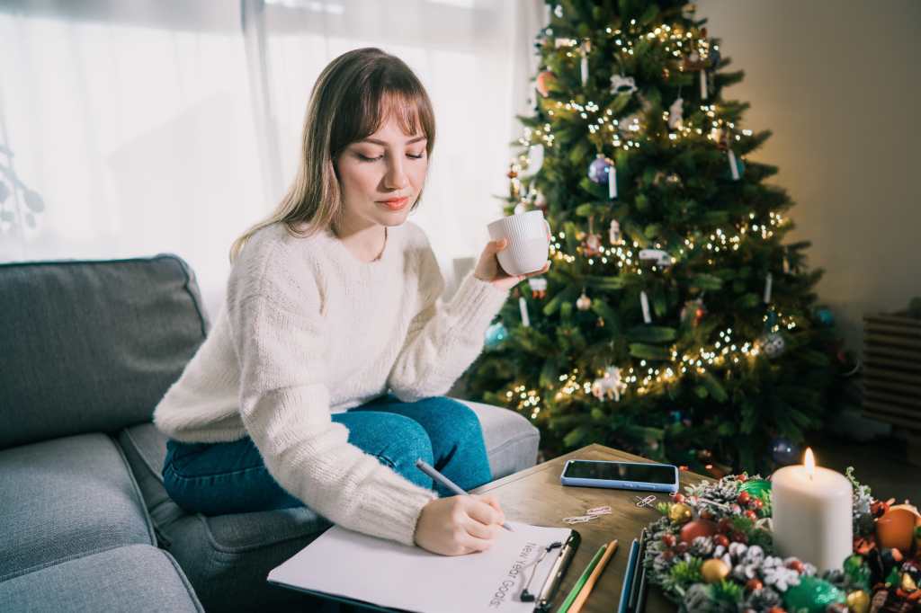Young Woman with cup (Mujer joven con taza) está escribiendo a mano los objetivos de Año Nuevo en un papel de nota en el día de Año Nuevo. Lista de resoluciones de año nuevo. Objetivos, resoluciones, planes, acción, concepto de lista de verificación. Plantilla de Año Nuevo, espacio de copia