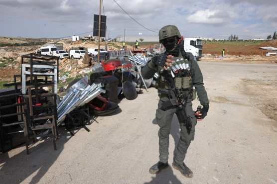 Israeli security officer stands near merchandise as Palestinians (unseen) recover what they can from pre-fabricated structures used as a shops and which are being removed by the Israeli military, at the entrance of the Israeli-occupied Palestinian West Bank village of al-Samu, close to the city of Hebron, on New Year's eve December 31, 2025. Israel has occupied the West Bank since 1967, and violence there has soared since the Gaza war erupted in October 2023 following Hamas's attack on Israel. (Photo by HAZEM BADER / AFP)
