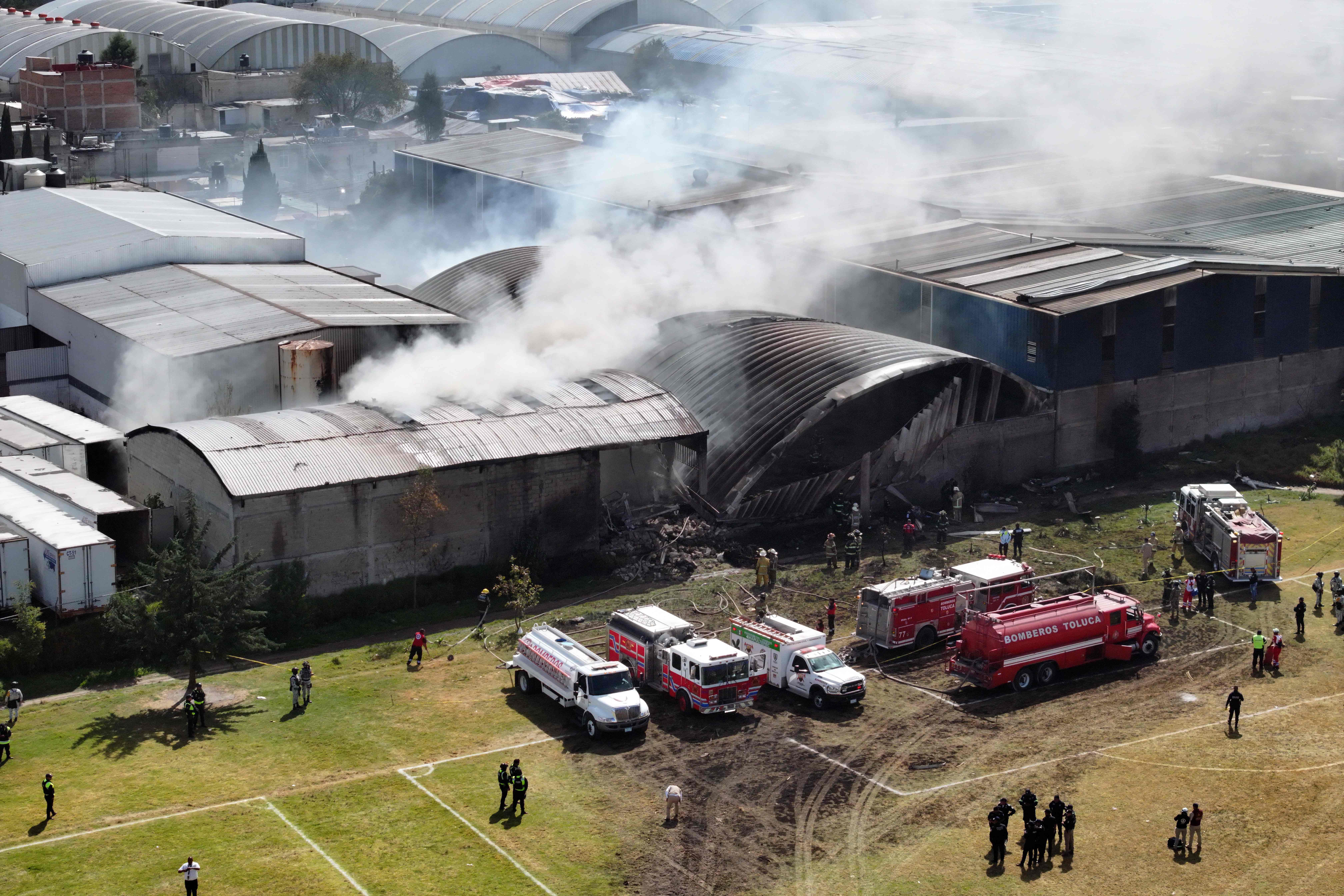 Aerial view of emergency services working to extinguish a fire caused by the crash of a Cessna Citation aircraft that struck an industrial warehouse in the Santa Maria Totoltepec neighborhood near Toluca International Airport in Mexico state on December 15, 2025. A small plane crashed shortly before reaching the airport in the central Mexican city of Toluca, killing at least four people, local Red Cross and authorities said. (Photo by MARIO VAZQUEZ / AFP)