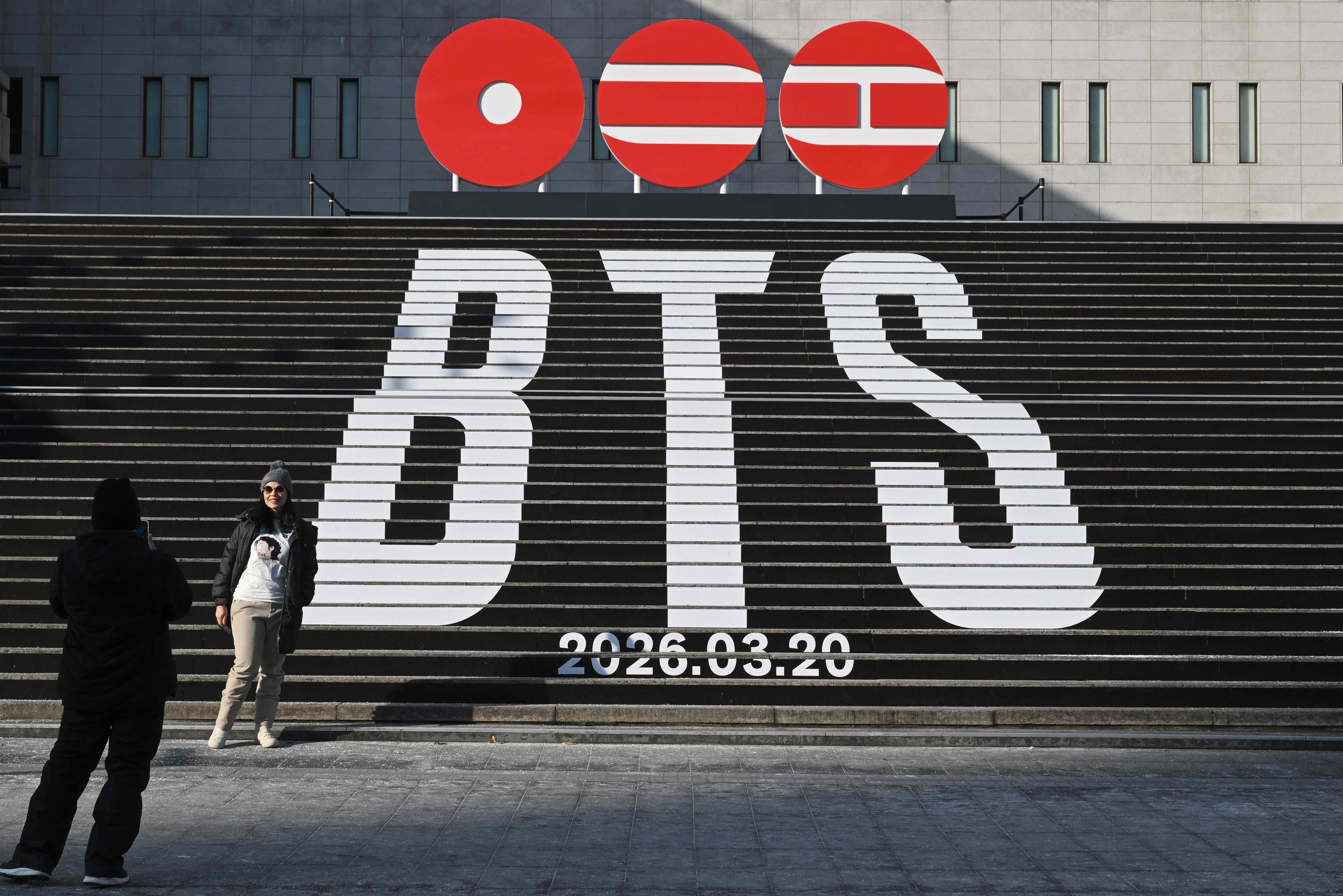 A woman poses for a photo in front of the stairs displayed with the logo of BTS' 2026 album and release date at Gwanghwamun Square in Seoul on January 14, 2026. K-pop megastars BTS will launch a comeback world tour from April, the band's label said on January 14. (Photo by Jung Yeon-je / AFP)