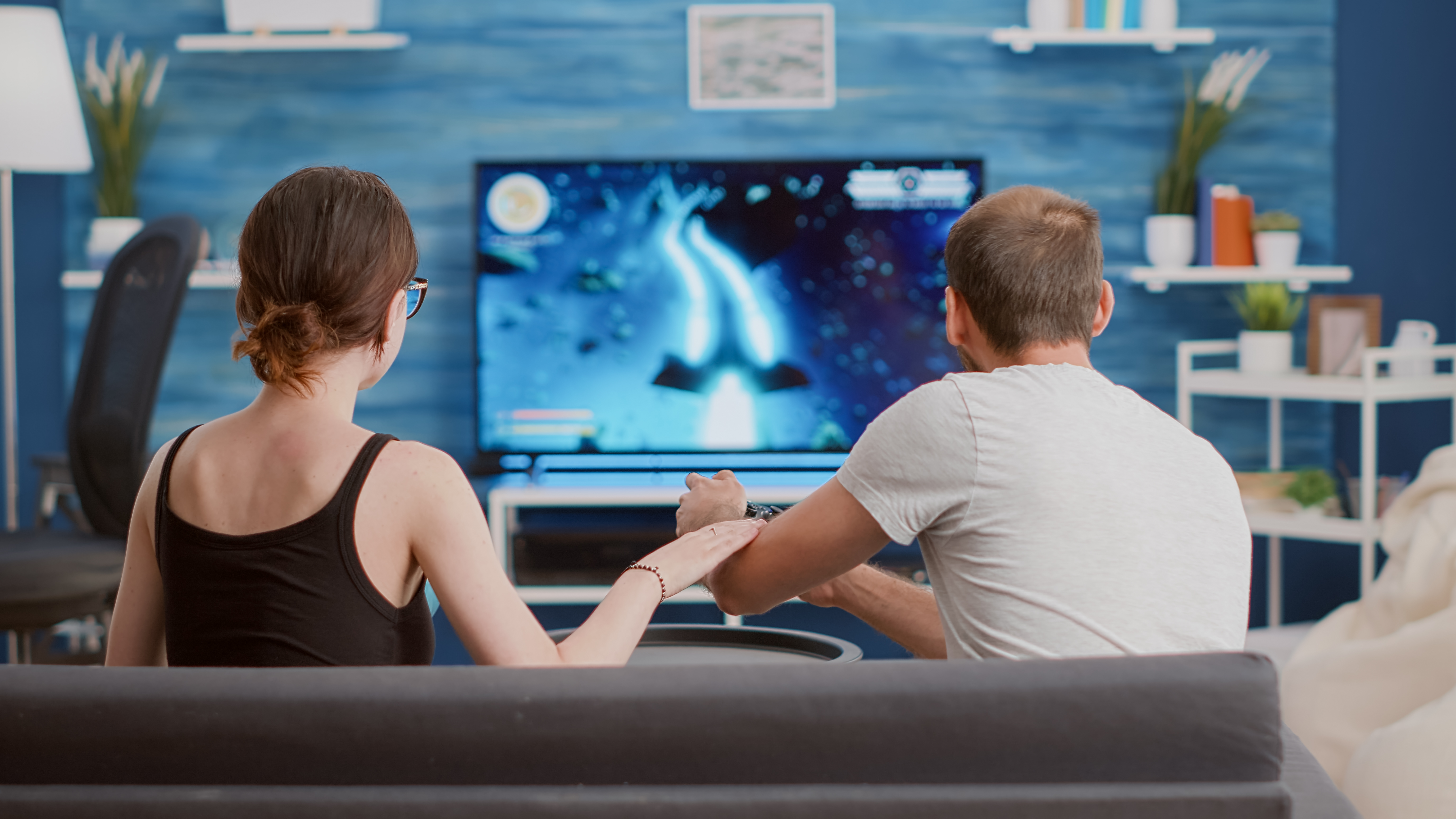 Girlfriend eating popcorn and supporting boyfriend gamer playing console video game first person shooter on tv while sitting on sofa. Couple on couch enjoying gaming simulation in modern living room.