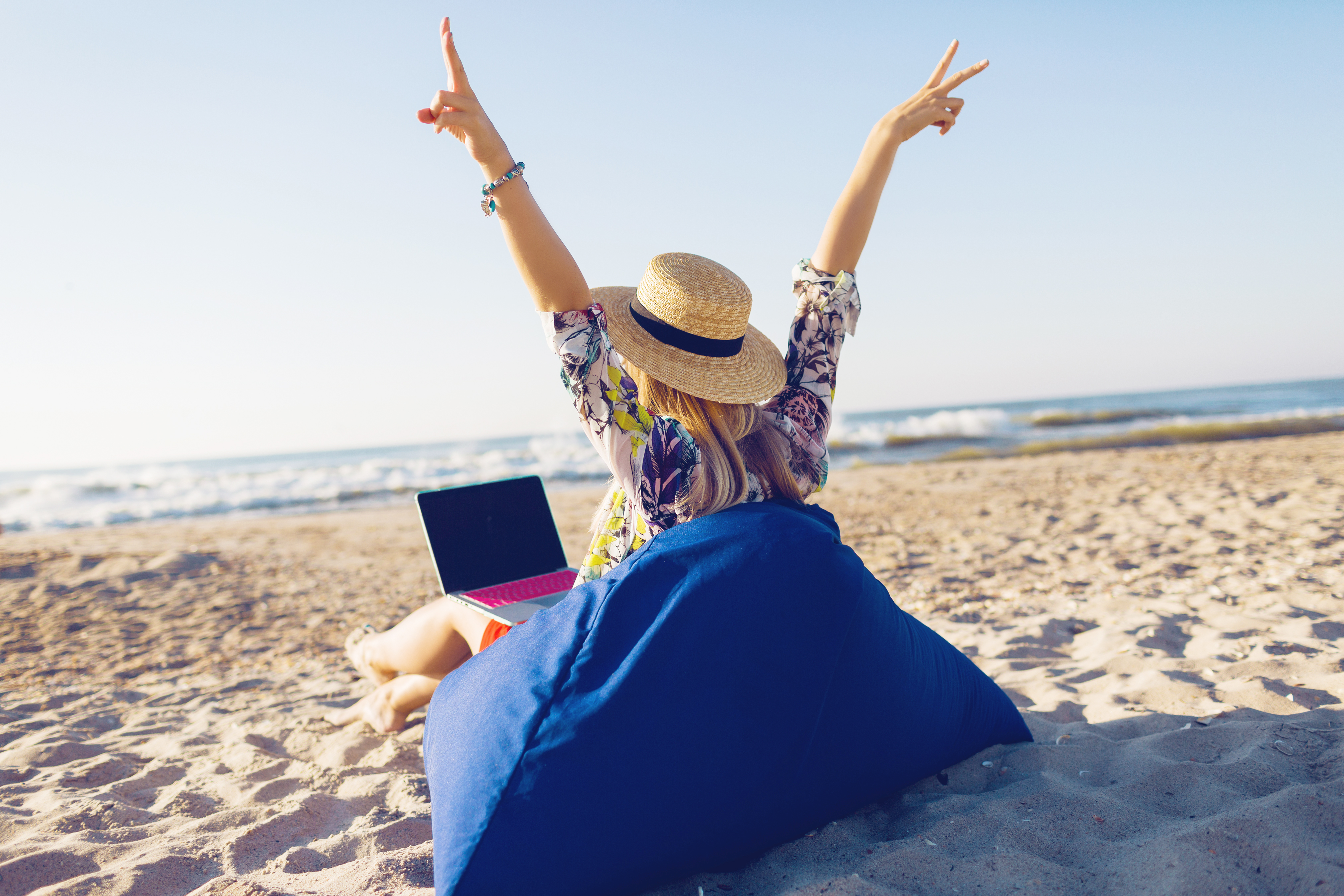 Beautiful young woman working with laptop on the tropical beach. Happy traveling woman in straw hat using laptop on holidays.