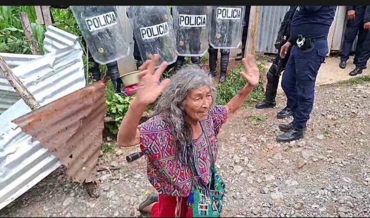 Tras el desalojo de su vivienda, Juana Santiago Chel permanece sentada frente a agentes de la PNC en Nebaj, Quiché. (Foto Prensa Libre: Captura de video)