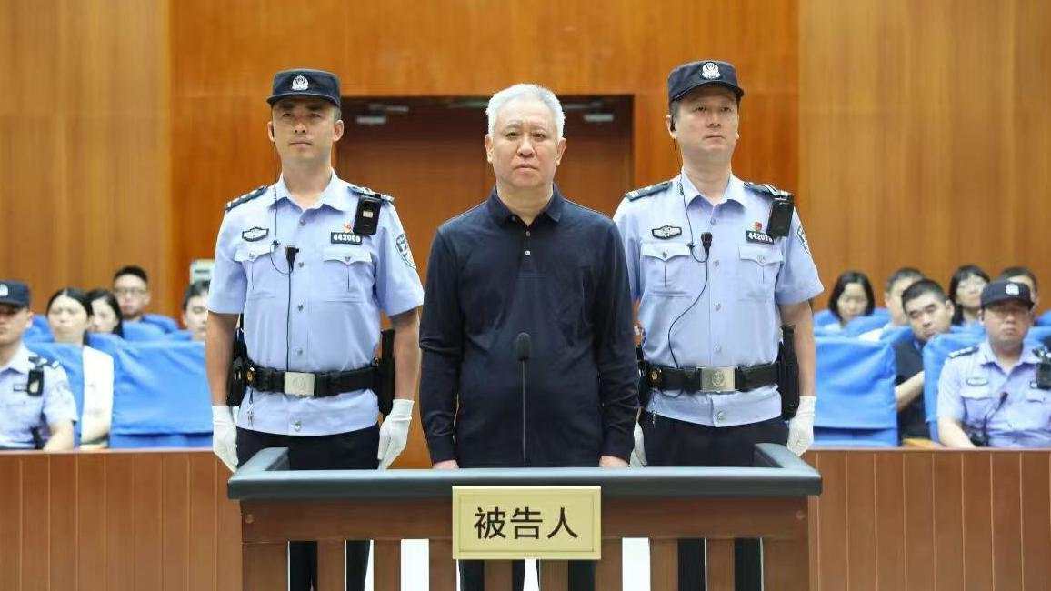 El fundador de China Evergrande Group, Hui Ka Yan, sube al estrado en un tribunal de China. Con expresión impasible, se encuentra entre dos agentes, con miembros del público al fondo.