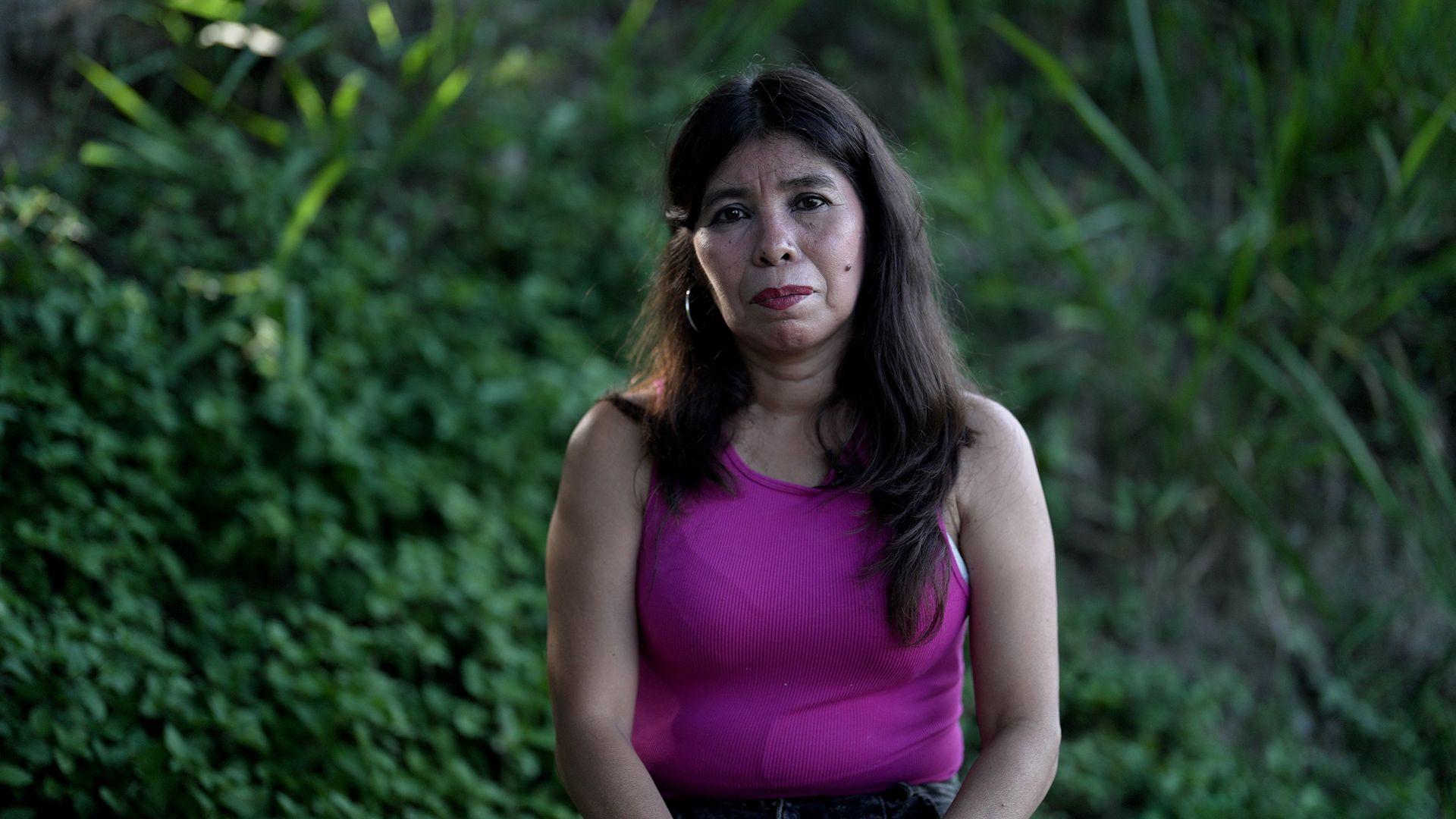 Una mujer sostiene el retrato de su hijo adulto. Está en un cuarto de una casa humilde, a media luz.