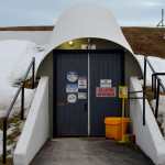 Dos puertas negras con señales de advertencia construidas en una colina cubierta de nieve, con algo de hierba seca asomando. Están cubiertas por un techo blanco en forma de arco y un camino, con paredes de hormigón blanco, está pavimentado frente a ellas.
