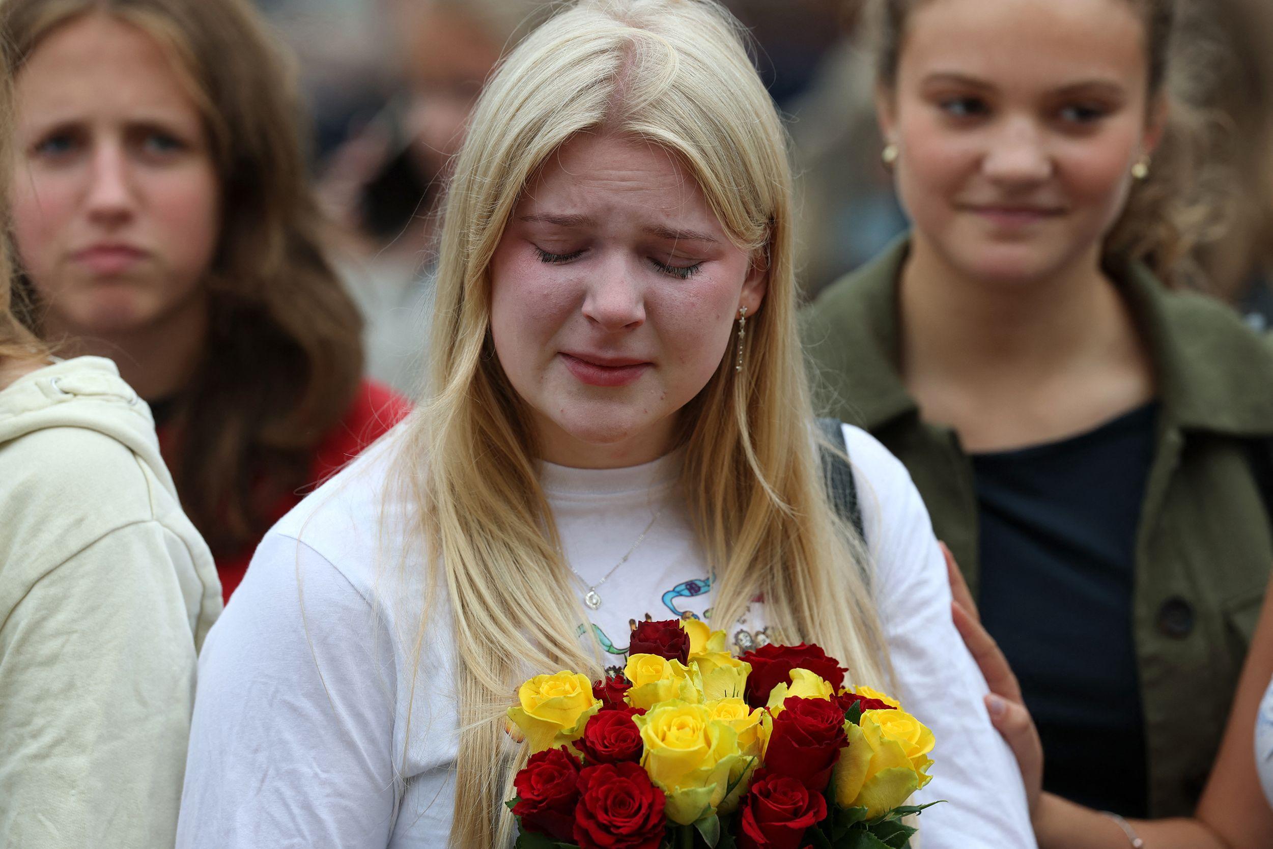 Personas observan una extensa ofrenda floral con numerosas banderas de Noruega, mientras un hombre y una niña permanecen junto a los homenajes.