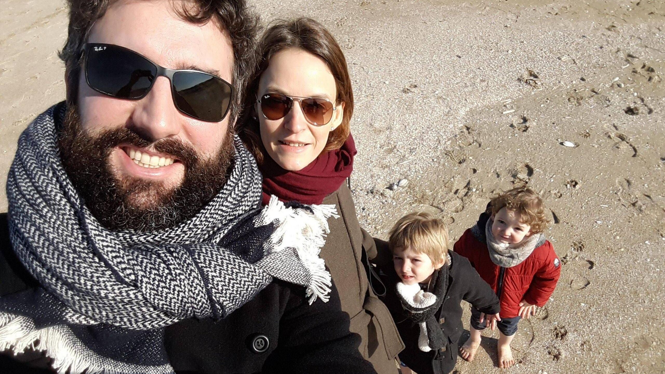Una fotografía en primer plano muestra a un hombre y una mujer, ambos de cabello castaño, posando juntos al aire libre y mirando a la cámara. El hombre lleva un sombrero blanco. Al fondo se aprecia una calle de la ciudad con la fachada de un edificio, mesas y sillas al aire libre y una moto naranja.