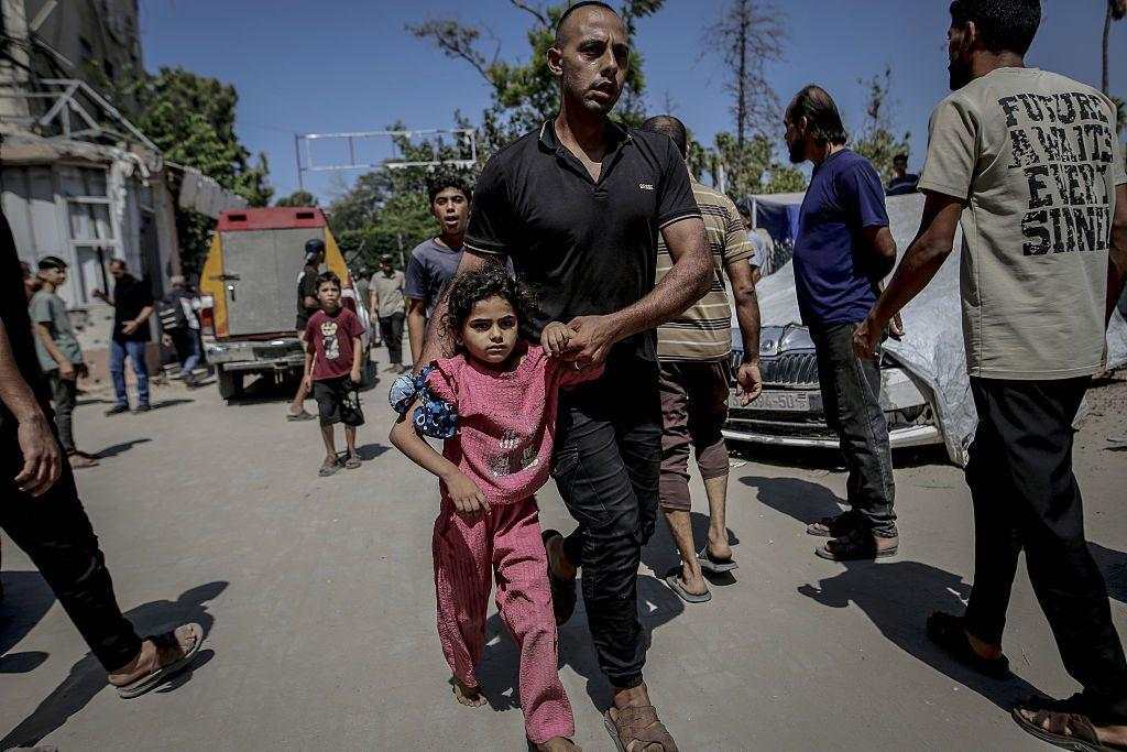 Hind Rajab sonríe a la cámara. Tiene el pelo largo, castaño y ondulado, y lleva una corona de flores rosas. También viste una camiseta rosa y una chaqueta negra con cremallera. Está sentada en una silla adornada con guirnaldas de flores rojas.