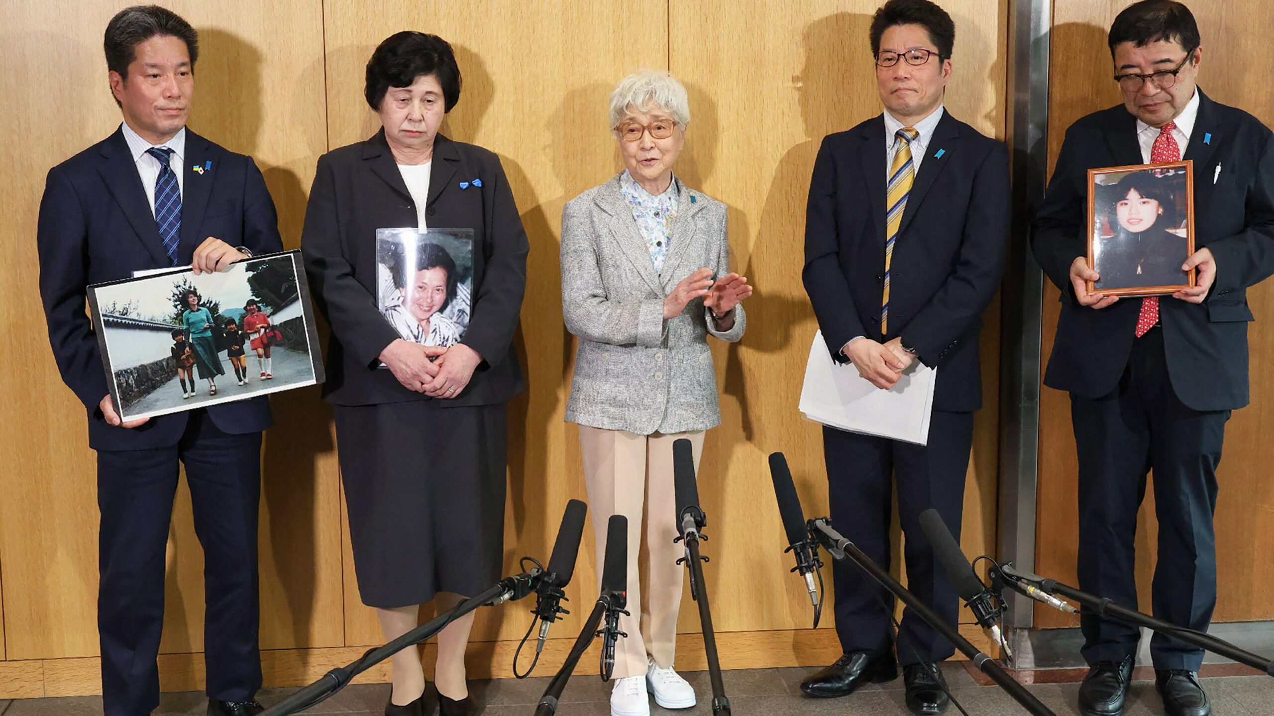Una foto antigua de Hitomi y su esposo, Charles. Están de pie frente a una fuente y un edificio de apartamentos. Hitomi lleva un blazer abotonado y una falda. Charles viste un traje gris con corbata a rayas. Hitomi tiene los brazos detrás de la espalda y Charles los tiene a los lados. Ambos sonríen y miran directamente a la cámara.