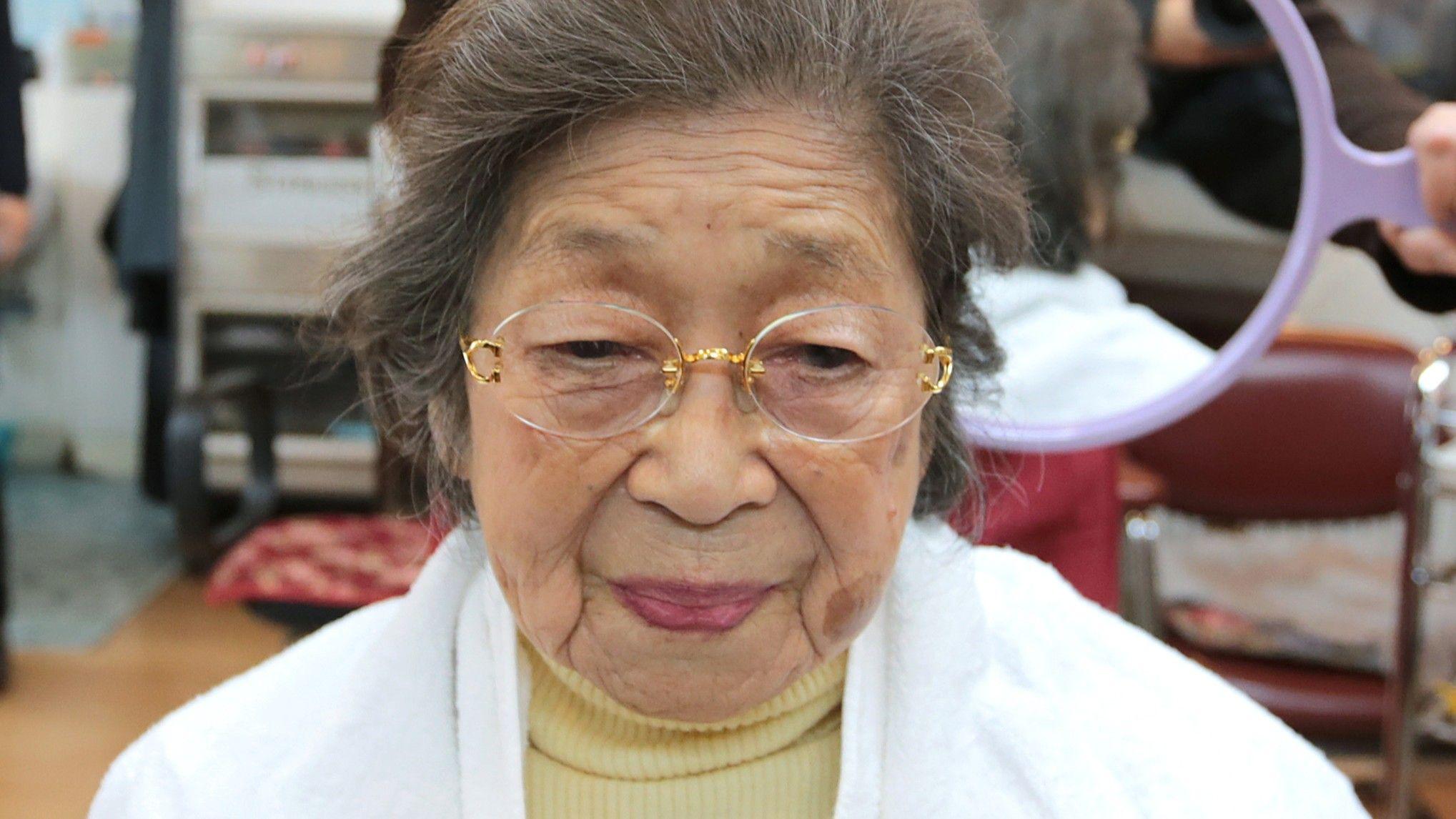 A 100-year-old Japanese woman wearing a butter yellow turtleneck, white jacket and rimless glasses at a hair salon with a hairdresser holding up a mirror behind her head