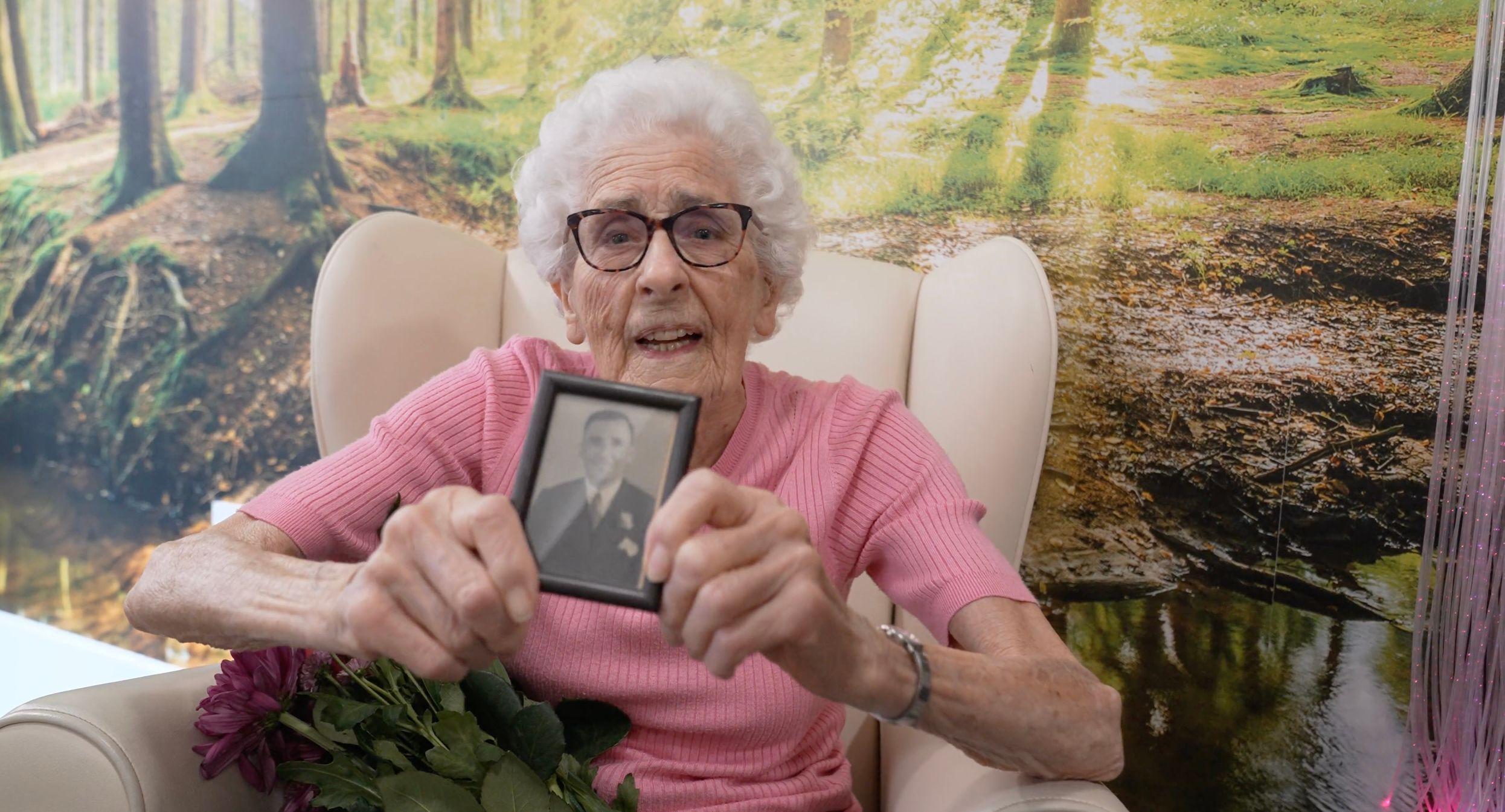 Una anciana con cabello blanco y gafas sostiene una fotografía antigua frente a la cámara y lleva una blusa rosa