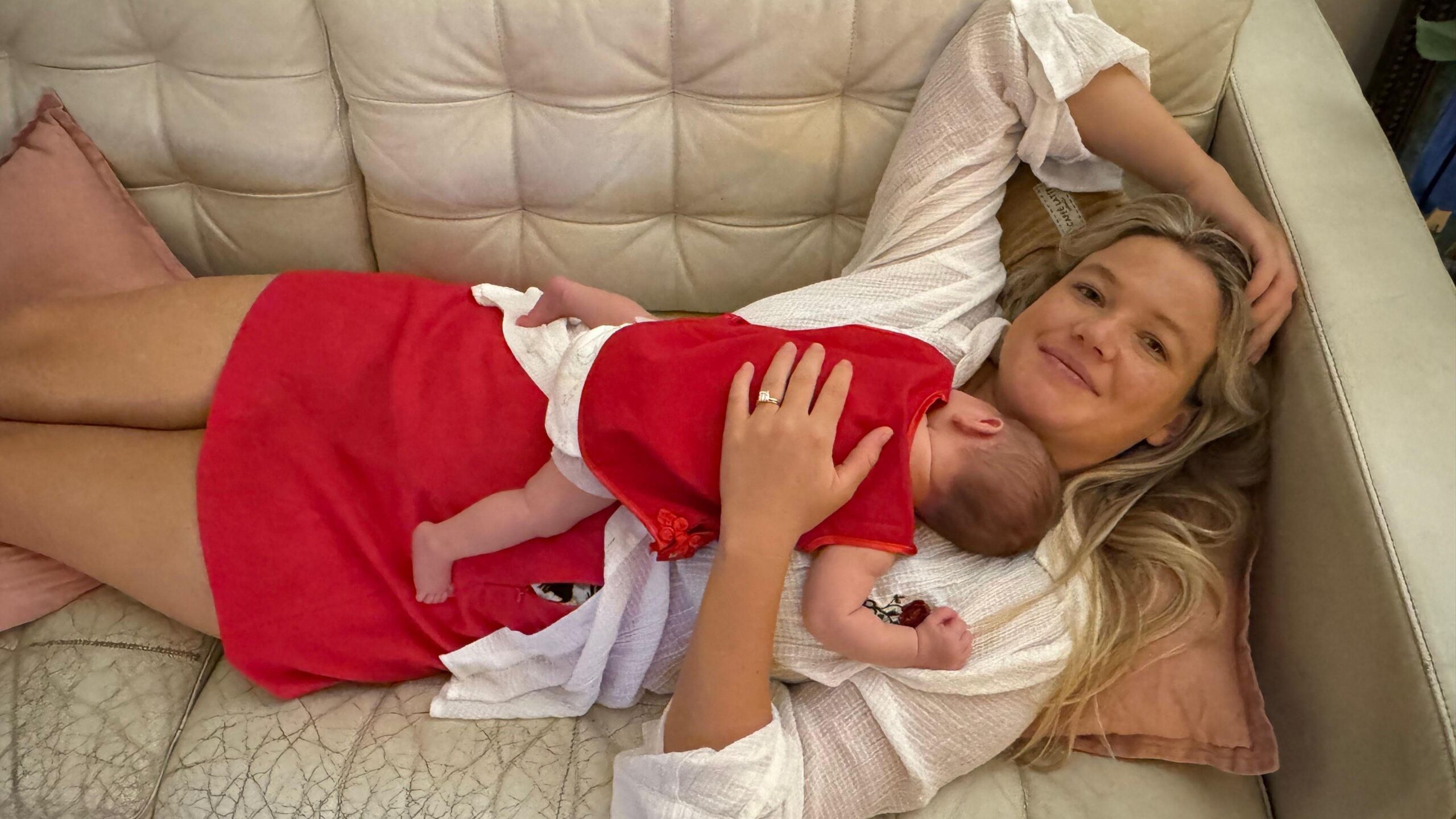 Una mujer de pelo largo y rubio, vestida con una blusa blanca y una falda roja, tumbada en un sofá color crema, con un bebé durmiendo sobre su pecho.