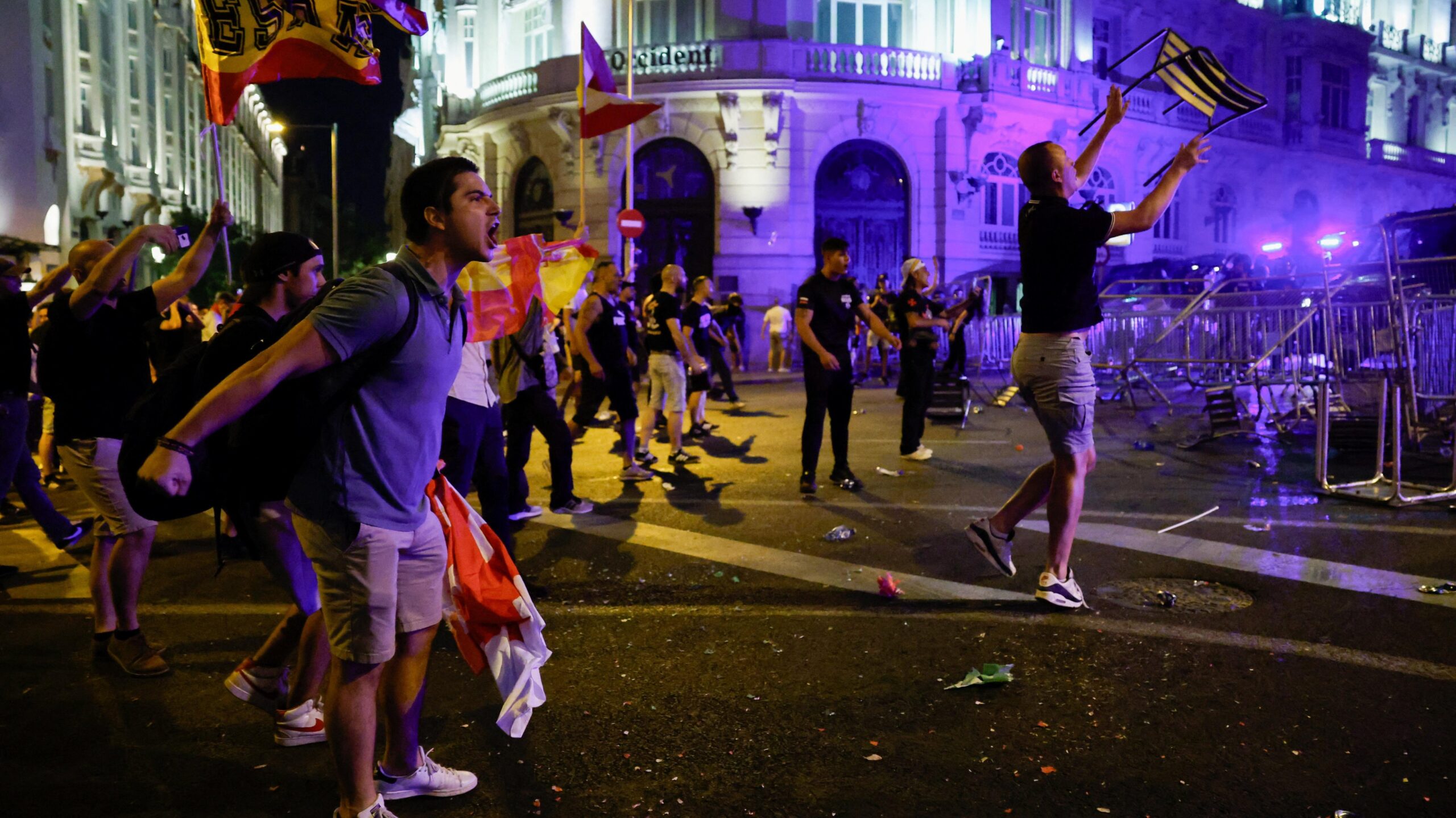 A woman at the front of a crowd chants with people behind and Spanish flags waving