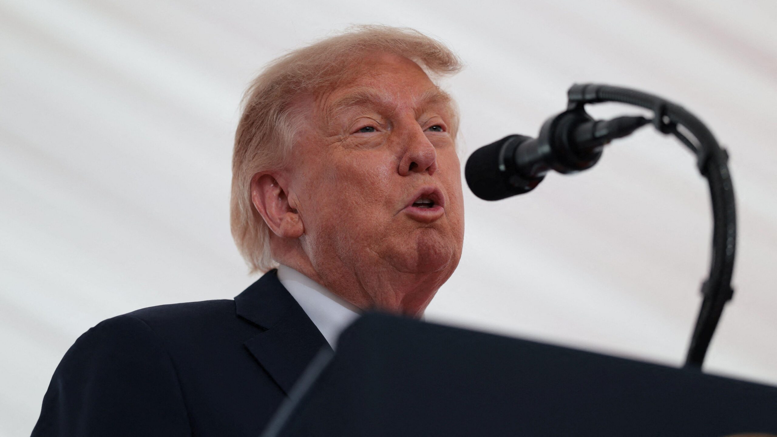 Trump está hablando por un micrófono. Lleva un traje negro y una camisa blanca. Detrás de él hay un fondo blanco.