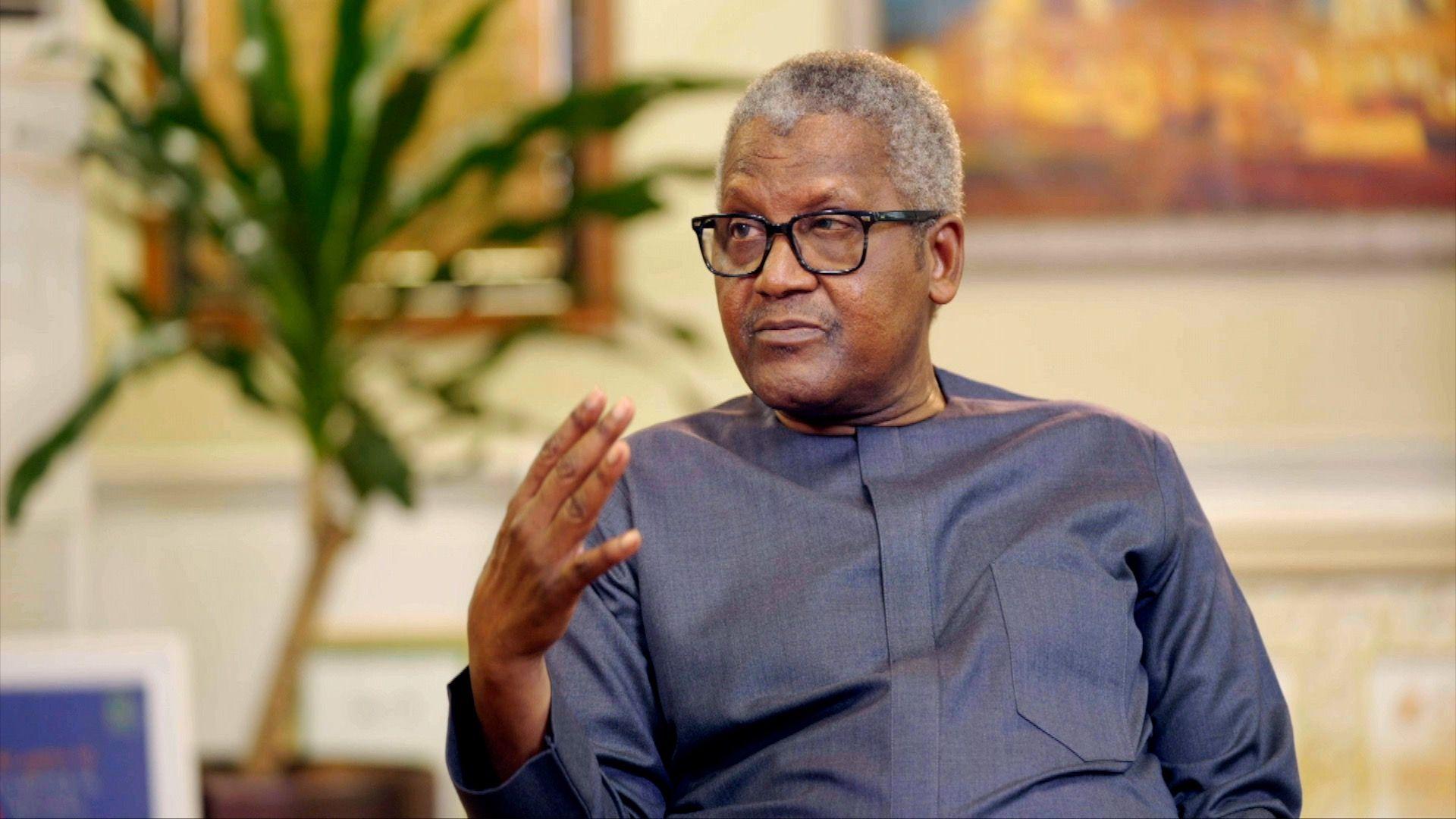 Esta imagen, retocada digitalmente, muestra un primer plano de Aliko Dangote, quien viste un traje negro, camisa blanca y corbata azul estampada. Detrás de él, se observa un rectángulo rojo, y sobre este, un fondo con la moneda nigeriana, el naira.