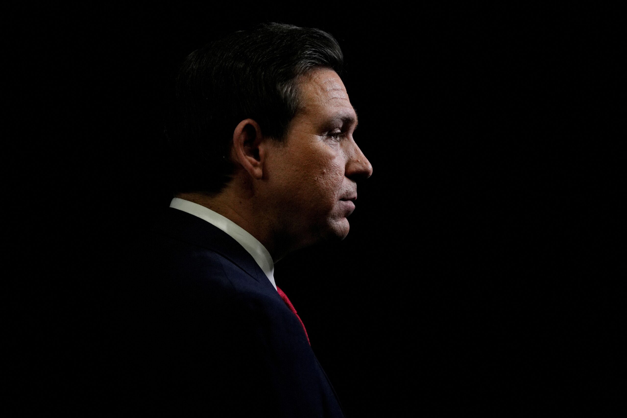 Ron DeSantis, en primer plano, vestido con traje oscuro y corbata. Está mirando hacia su derecha.