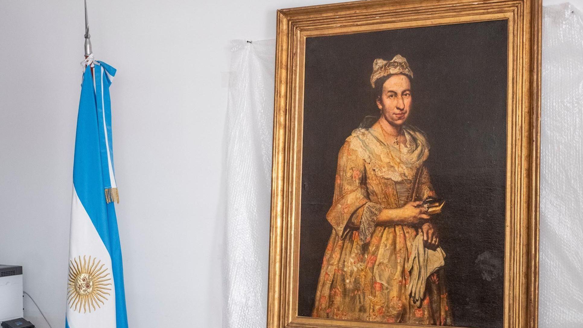 En la pared cuelga un retrato con marco dorado sobre fondo blanco, junto a él hay una bandera argentina enrollada.