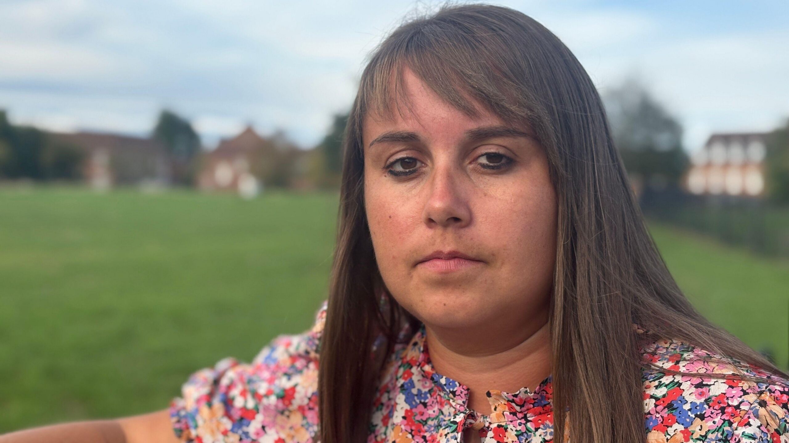 Becky Jones tiene el pelo castaño hasta los hombros con flequillo. Está de pie en un parque y lleva un vestido con flores multicolores.