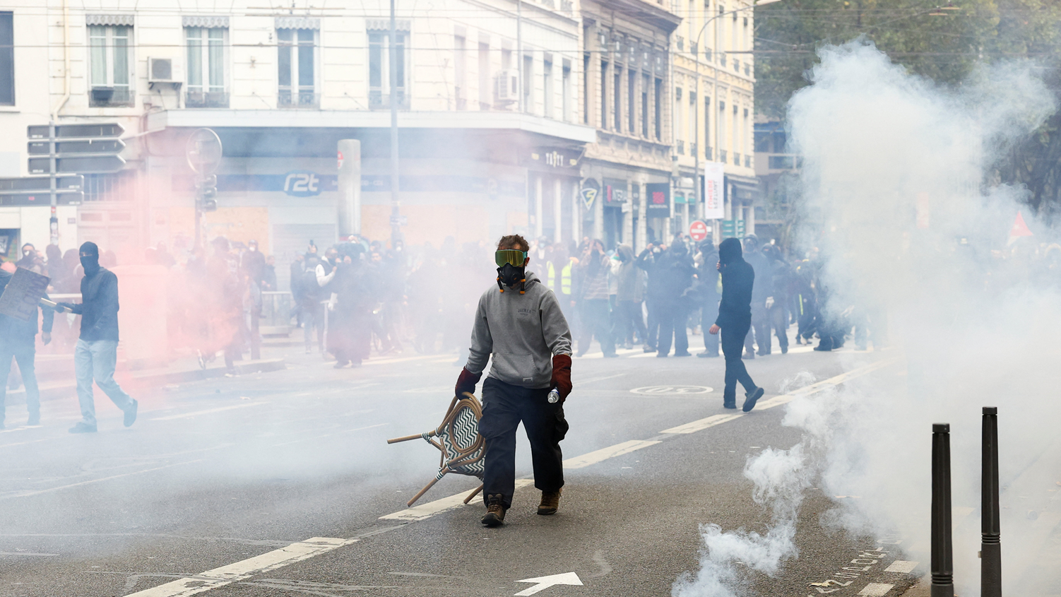 Un manifestante, con una máscara antigás y unos lentes especiales, arrastra una silla en medio del humo provocado por los gases lacrimógenos en una calle. Atrás se ven varias personas.