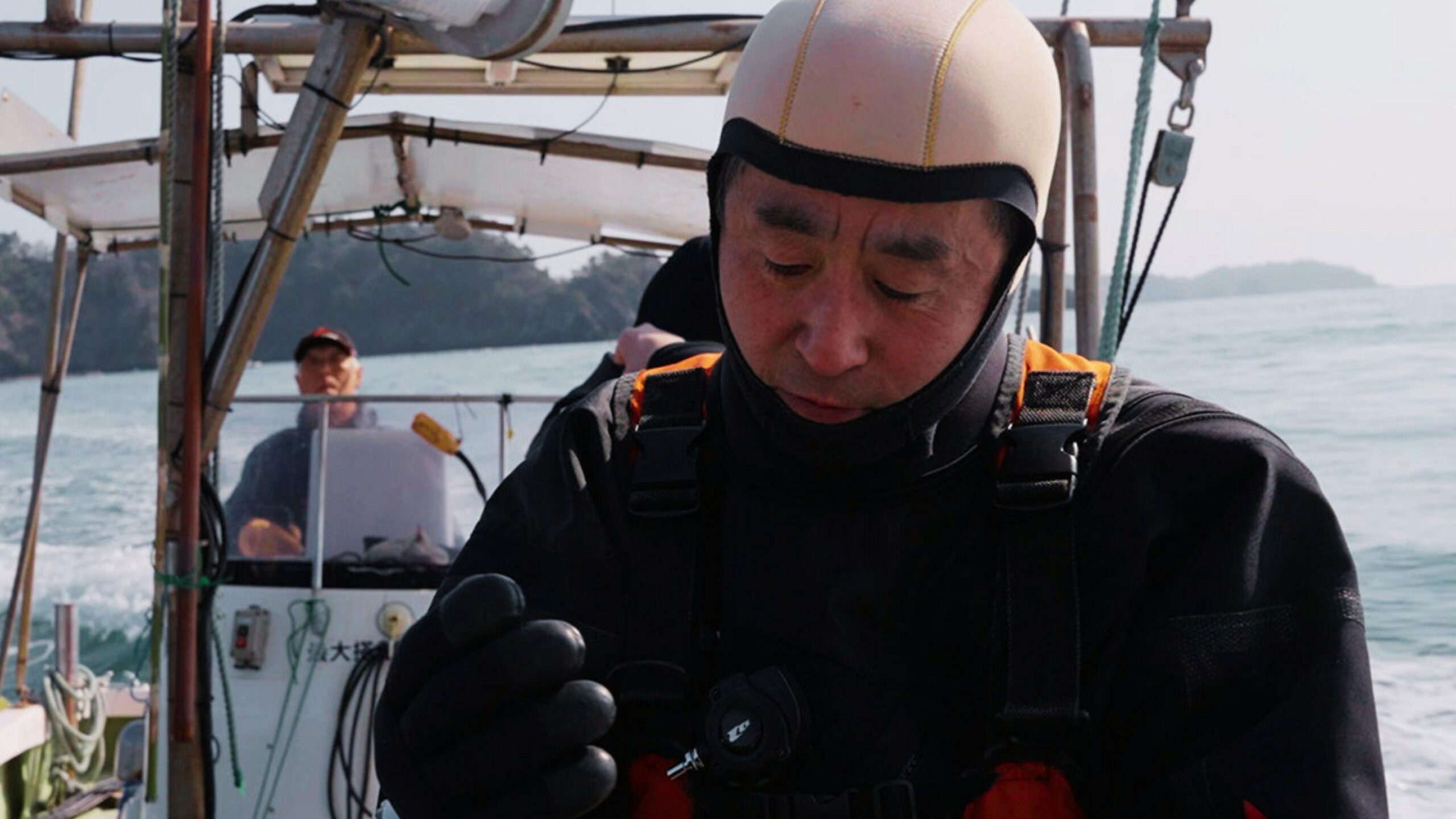 Un hombre (Yasuo), vestido con un traje de neopreno negro, guantes de neopreno y una capucha de buceo de color claro, aparece en primer plano desde una pequeña embarcación, mirando hacia abajo mientras manipula el equipo de buceo. La embarcación navega en aguas tranquilas con una costa boscosa al fondo, y se observa a otra persona al timón detrás de un parabrisas; un pequeño texto en caracteres japoneses aparece en la consola de la embarcación.