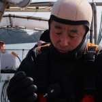 Un hombre (Yasuo), vestido con un traje de neopreno negro, guantes de neopreno y una capucha de buceo de color claro, aparece en primer plano desde una pequeña embarcación, mirando hacia abajo mientras manipula el equipo de buceo. La embarcación navega en aguas tranquilas con una costa boscosa al fondo, y se observa a otra persona al timón detrás de un parabrisas; un pequeño texto en caracteres japoneses aparece en la consola de la embarcación.