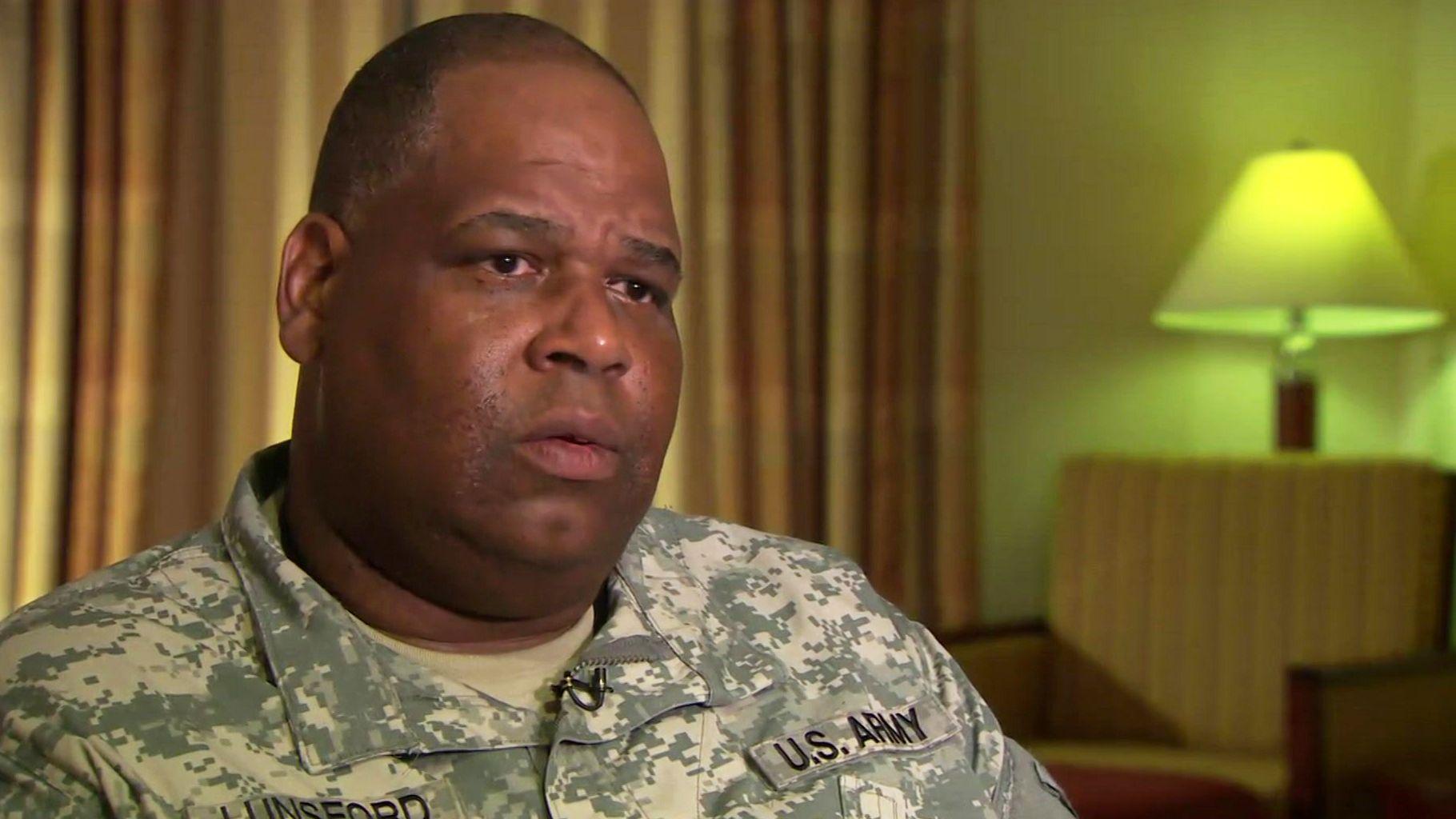 Lunsford is on the left side of the photo - a crop of his head and shoulders are visible. He is wearing military fatigues. Behind him are drawn light brown curtains and a table lamp