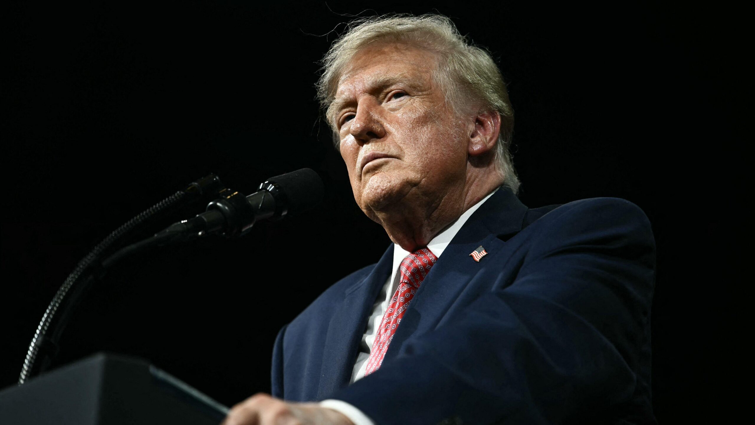 El presidente Trump aparece de pie frente a un podio negro. Viste una camisa blanca, una chaqueta azul con un pin de la bandera estadounidense y una corbata roja estampada. No sonríe y sujeta los bordes del podio. La fotografía lo muestra desde abajo, mirando hacia arriba.