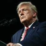 El presidente Trump aparece de pie frente a un podio negro. Viste una camisa blanca, una chaqueta azul con un pin de la bandera estadounidense y una corbata roja estampada. No sonríe y sujeta los bordes del podio. La fotografía lo muestra desde abajo, mirando hacia arriba.